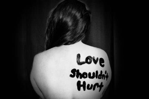 A white woman with dark hair facing away from the camera to a black background. Written on the right (to us) side of her naked back in thick black is "Love Shouldn't Hurt"