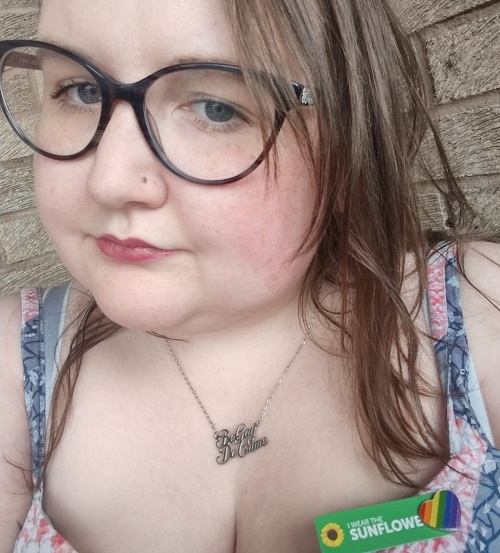 Caroline - white woman with brown hair and glasses, Pinned to her dress with a rainbow heart pin badge is a Sunflower Hidden Disabilities key fob. She is not wearing a mask
