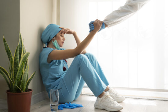 a stressed nurse sits on the floor holding her head, a hand reaches out to help her up