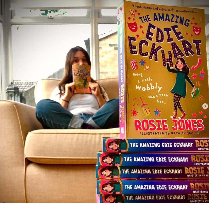 Author Rosie Jones sat on a sofa in the background, with a stack of copies of The Amazing Edie Eckhart in the foreground