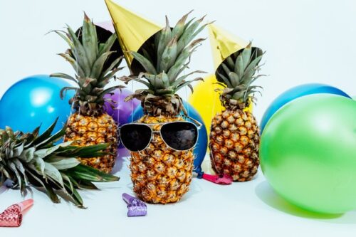 Three pineapples, surrounded by balloons and party blowers, one is "wearing" sunglasses