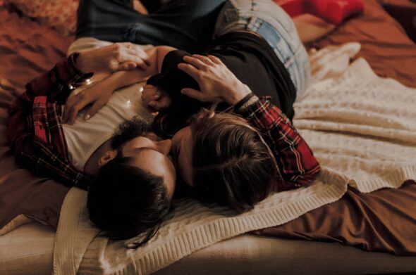 A couple lying on a bed, embracing. One person is masculine presenting in a checked shirt with a beard and brown hair, the other has brown hair and may be wearing make up and nail polish
