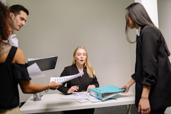 A feminine presenting person with long blonde hair is sat at a desk looking overwhelmed as three other people place files and folders in front of her