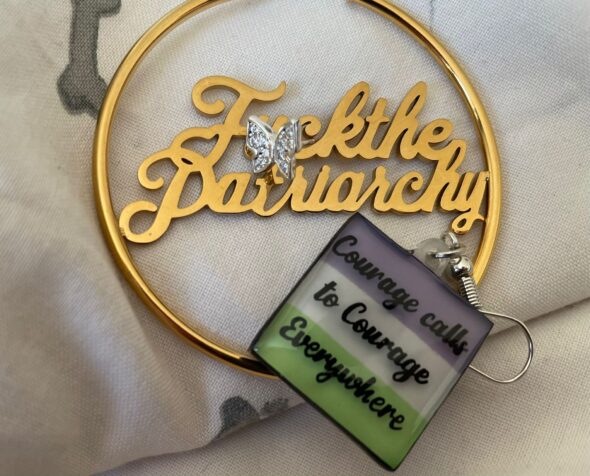 Protest jewelery, a gold hoop with "F*ck the Patriarchy" and an earring in suffragette colours (purple, white and green) which reads "courage calls to courage everywhere"