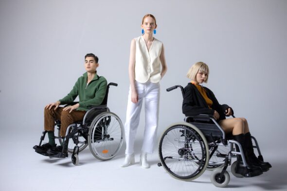 Three people - two using wheelchairs - one of them is male presenting with dark hair, the other female presenting with blonde hair. The person standing is female presenting with red tied back hair and a hand in her pocket
