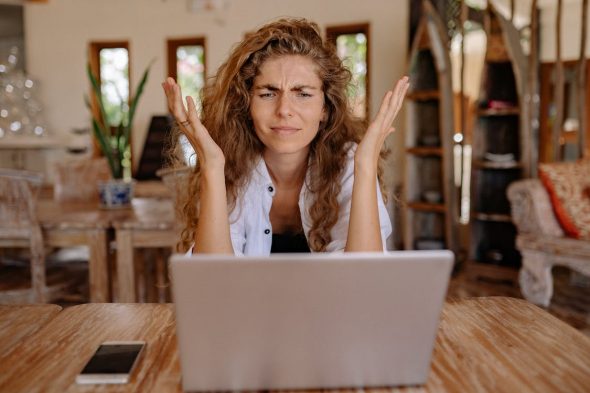 Feminine presenting person with brown hair in front of her laptop with her hands lifted up in exasperation