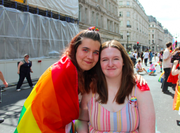 Shona and her partner - two femme presenting people with brown hair, one is wrapped in a rainbow flag and Shona is in a wheelchair
