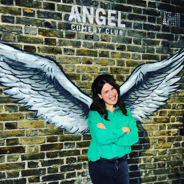 Comedian Rosie Jones outside Angel Comedy Club - in front of wing graffiti. She is feminine presenting with dark hair, in a green top with her arms crossed
