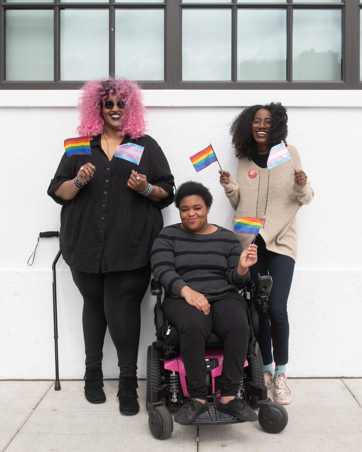 Three Black and disabled folx smile and hold mini flags. On the left, a non-binary person holds both a rainbow pride flag and a transgender pride flag, while a cane rests behind her. In the middle, a non-binary person waves the rainbow flag while in their power wheelchair. On the right, a femme waves both a rainbow and transgender pride flag.